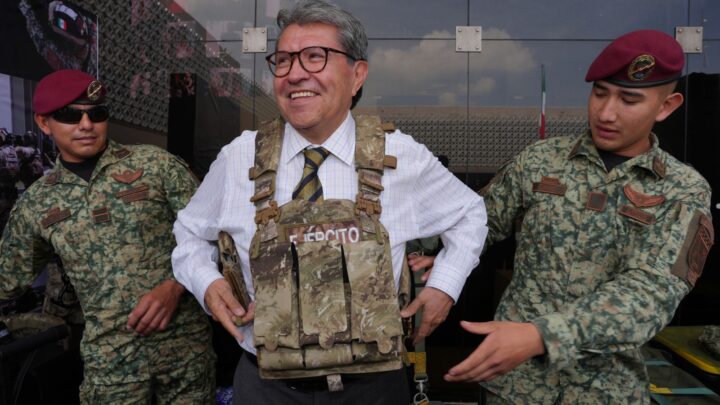 Ricardo Monreal prueba un chaleco táctico acompañado de dos militares durante una exhibición.